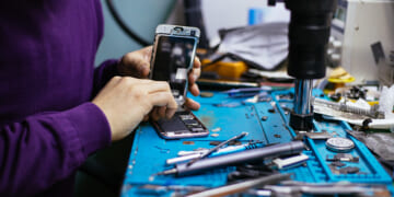 Technician repairing mobile phone at a station with microscope, heat gun, and blue mat